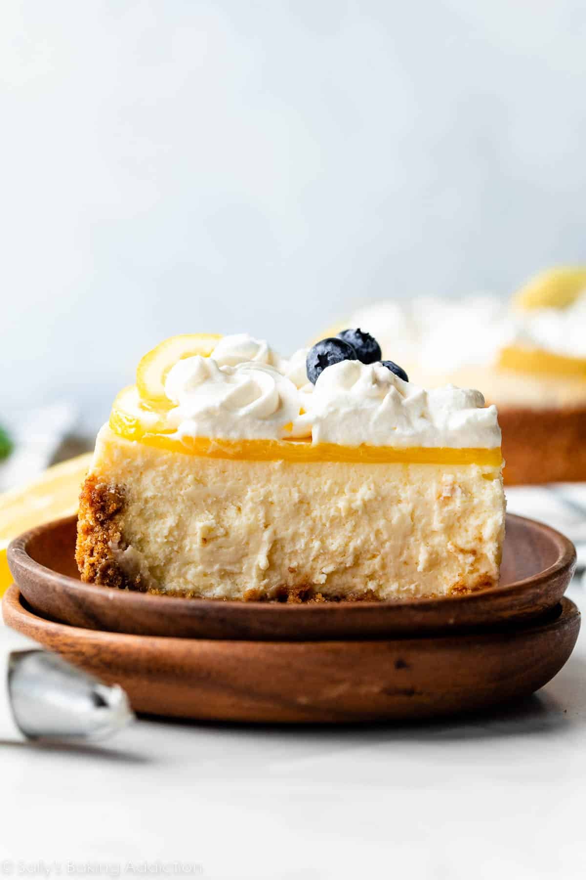 slice of lemon cheesecake with lemon curd and whipped cream on brown wooden plate.
