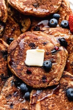 close-up of whole wheat blueberry pancakes with maple syrup, butter, and fresh blueberries on top.