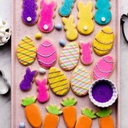 decorated Easter cookies on pink baking sheet including bunnies with marshmallow tails, Easter eggs, and carrots with orange and green icing.
