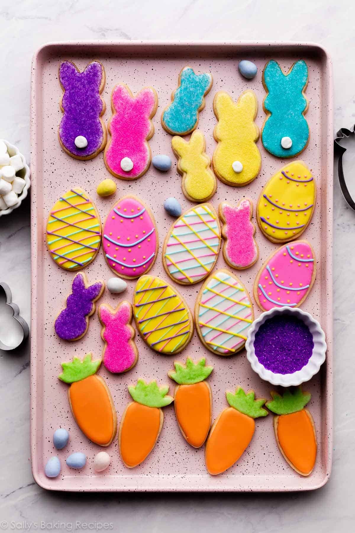 decorated Easter cookies on pink baking sheet including bunnies with marshmallow tails, Easter eggs, and carrots with orange and green icing.