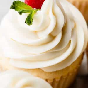 close-up photo of white chocolate frosting buttercream swirled on cupcake with raspberry and fresh mint garnish.