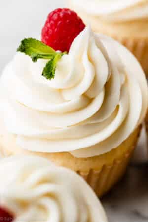 close-up photo of white chocolate frosting buttercream swirled on cupcake with raspberry and fresh mint garnish.