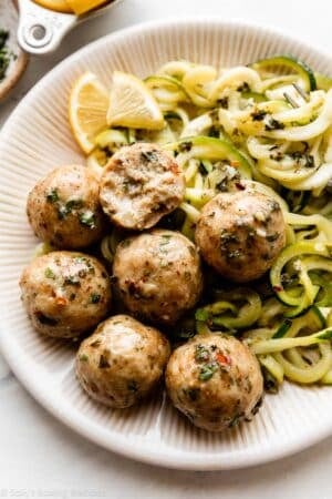 chicken meatballs with herbs on top on a plate with zucchini noodles and lemon slices.