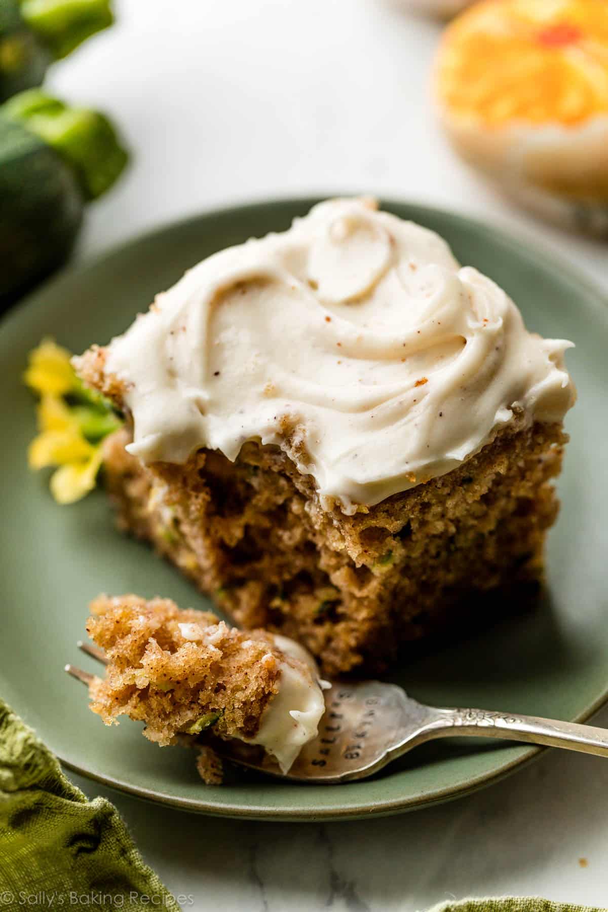 square slice of zucchini cake with cream cheese frosting on green plate with piece of the cake on a fork.