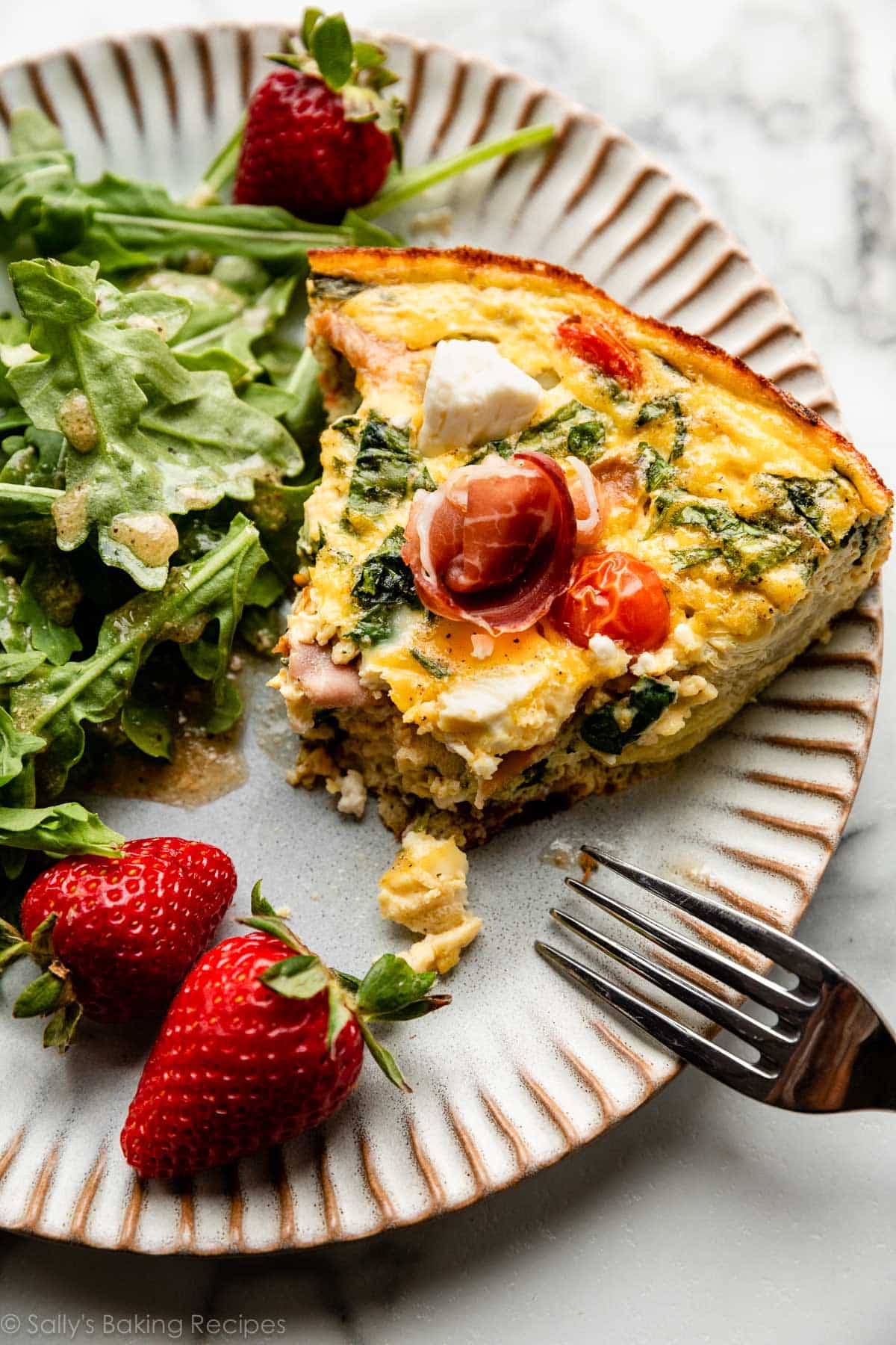 spinach, tomato, and feta cheese frittata slice on plate with arugula salad and strawberries.