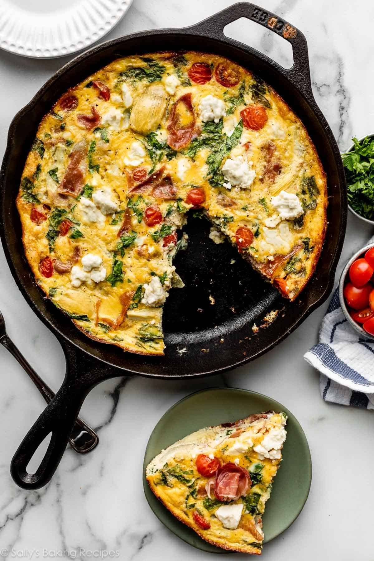 Mediterranean-inspired frittata in cast iron skillet with slice taken out and placed on green plate next to it.