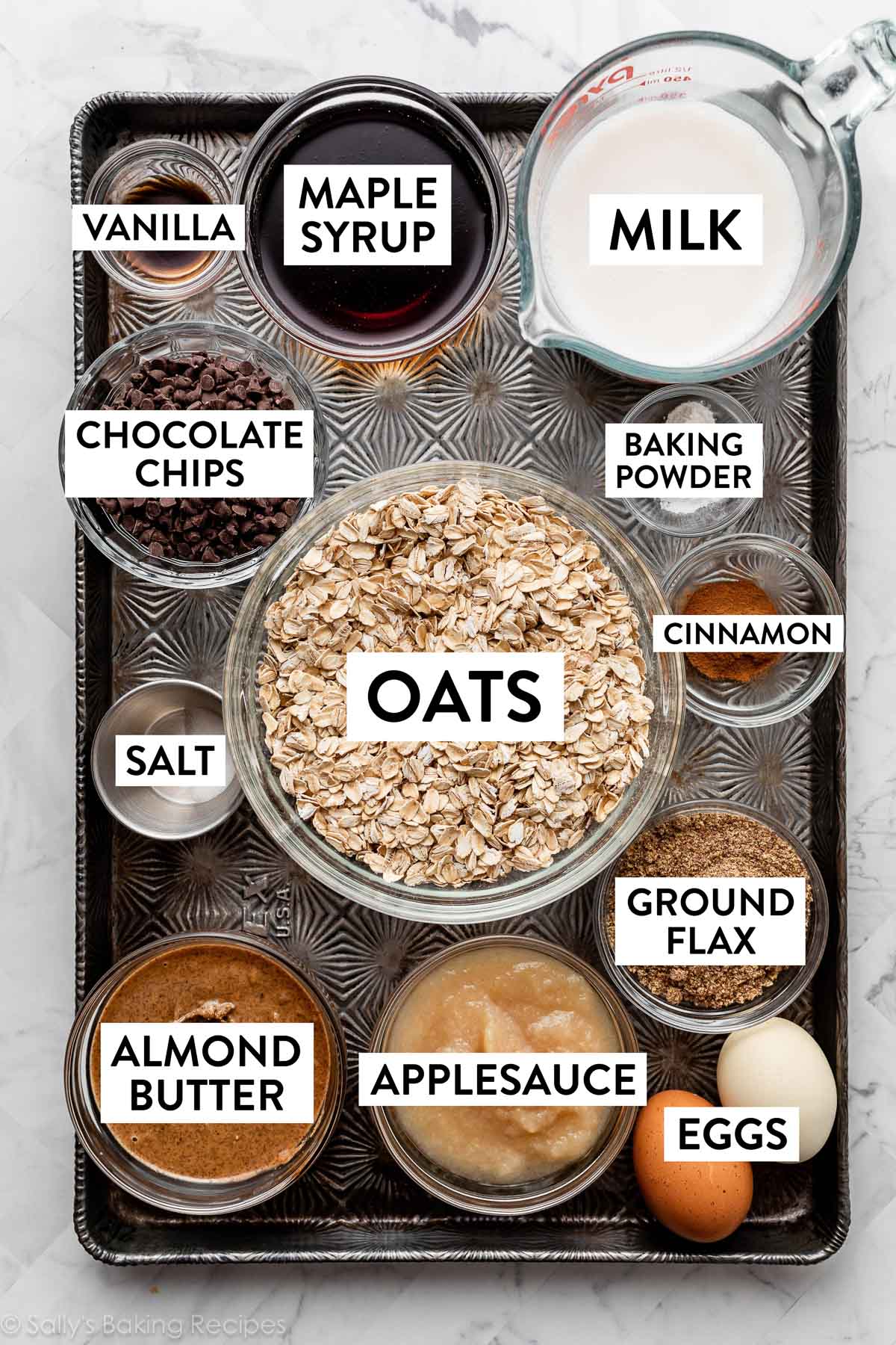 ingredients on baking sheet including milk, oats, ground flax, almond butter, applesauce, eggs, maple syrup, and more.