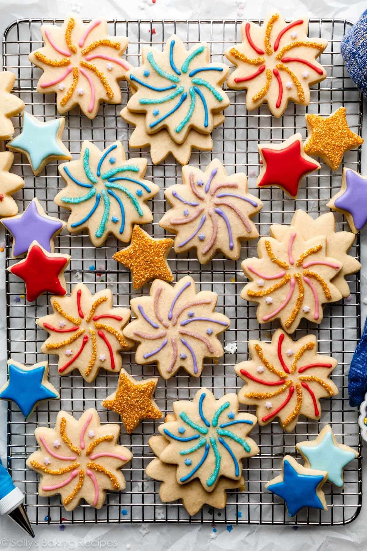 decorated fireworks celebration and star-shaped sugar cookies with various colors of icing and gold sprinkles on top of wire cooling rack.