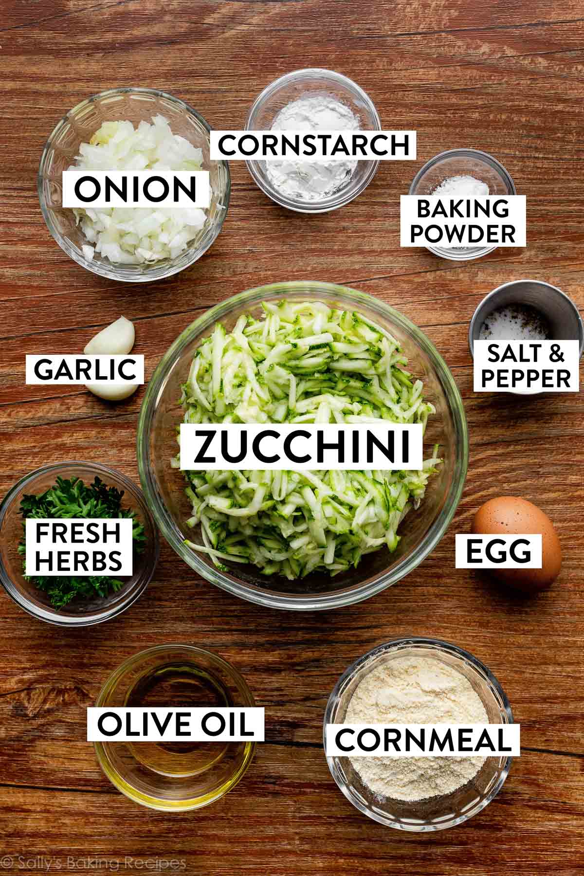 bowls of ingredients on wooden backdrop including shredded zucchini, chopped onion, cornstarch, cornmeal, baking powder, fresh herbs, egg, and garlic.