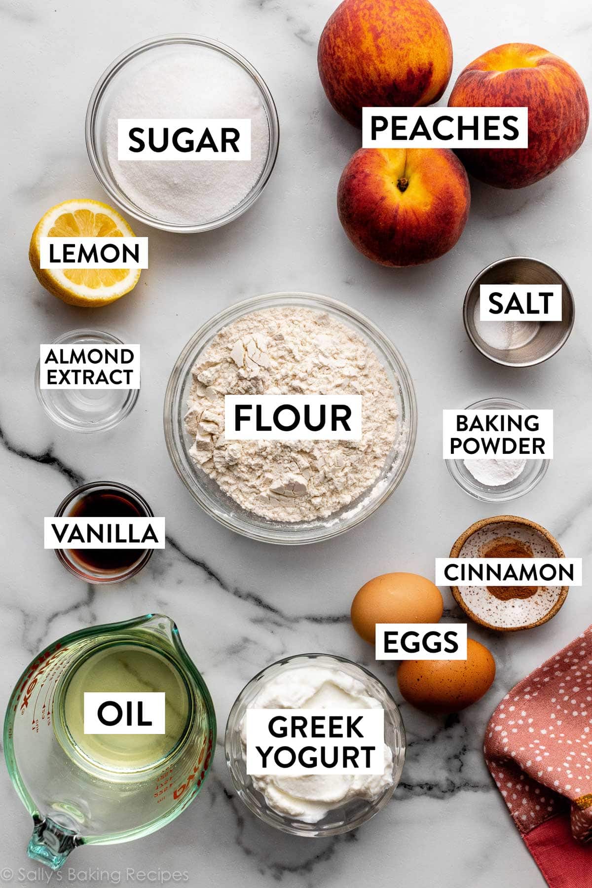 ingredients on marble counter including peaches, sugar, lemon, salt, flour, oil, greek yogurt, eggs, and others.