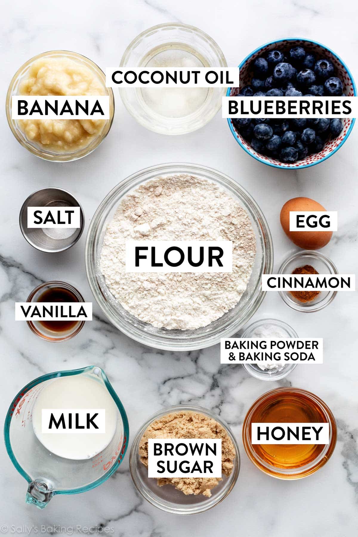 ingredients on counter including flour, blueberries, mashed banana, honey, milk, and coconut oil.