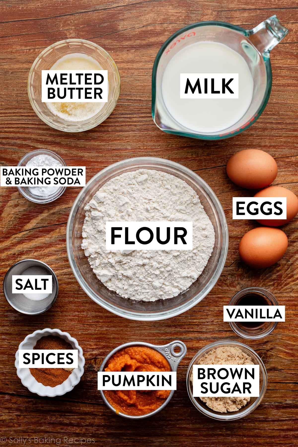 ingredients in bowls on a wooden surface including flour, milk, melted butter, brown sugar, pumpkin, spices, vanilla, salt and eggs.