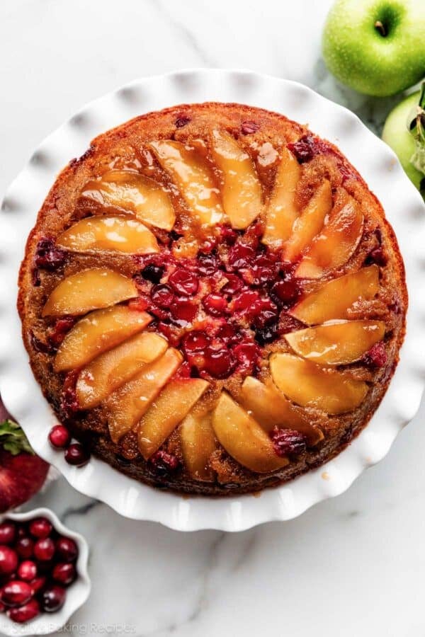 cranberry apple upside down fall cake on white cake stand with green apples next to it on the counter.