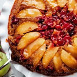 cranberry apple upside down fall cake on white cake stand with slice being removed.