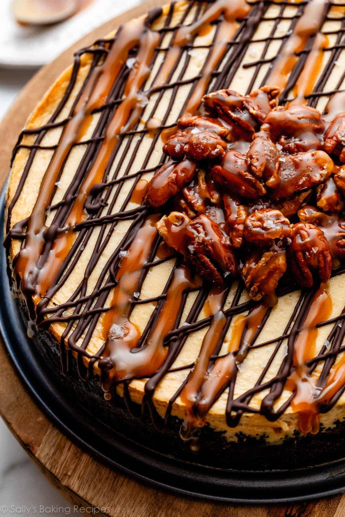 close up of turtle cheesecake with salted caramel sauce, melted chocolate, and pile of caramel pecans on top.