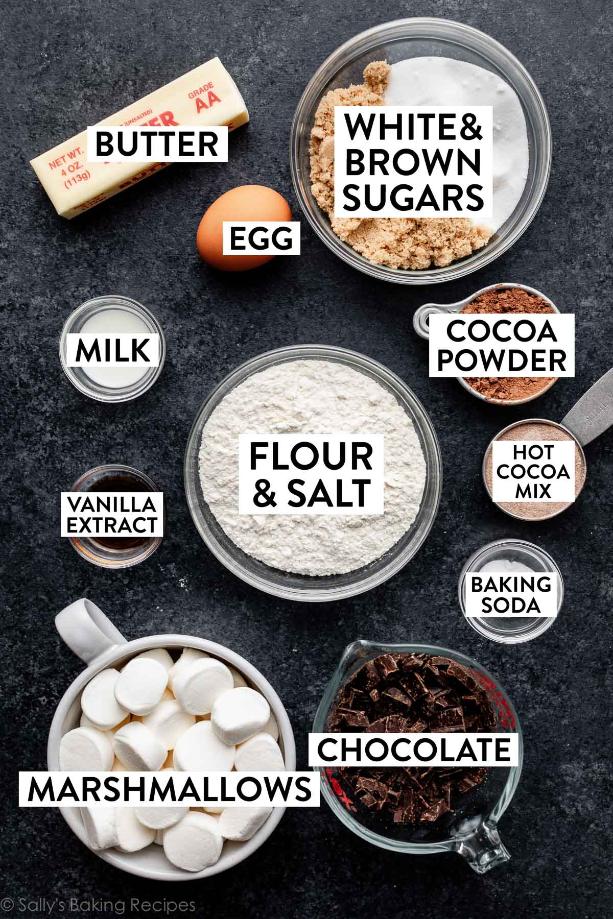 ingredients on black-ish blue-ish backdrop including butter, brown and white sugars, chocolate, flour, salt, cocoa, and hot cocoa mix.