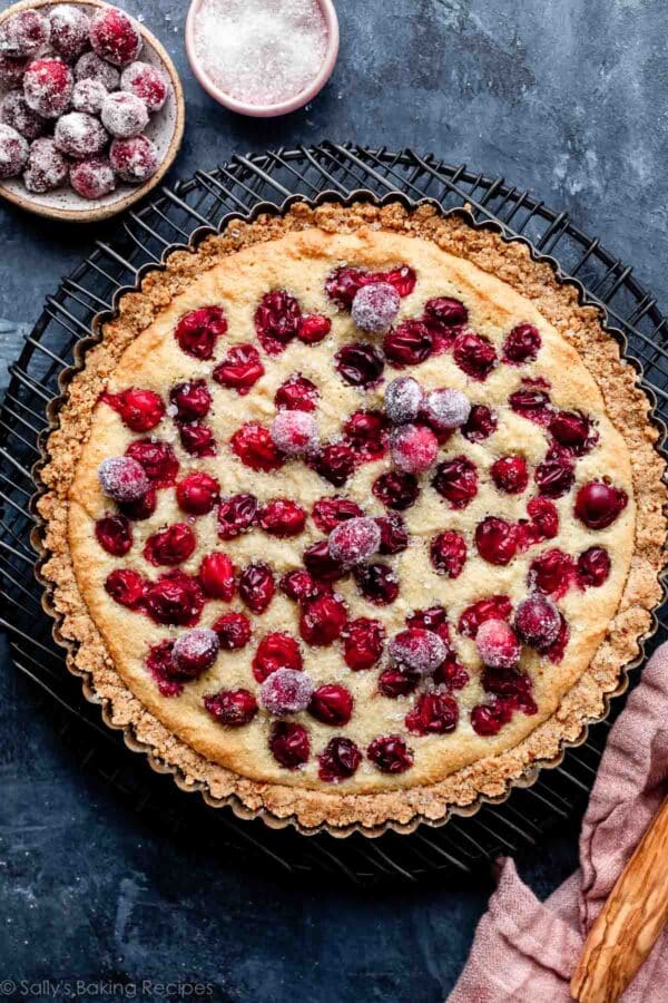 cranberry frangipane tart with almond crust and sugared cranberries on top of a round black wire rack on a blue surface.