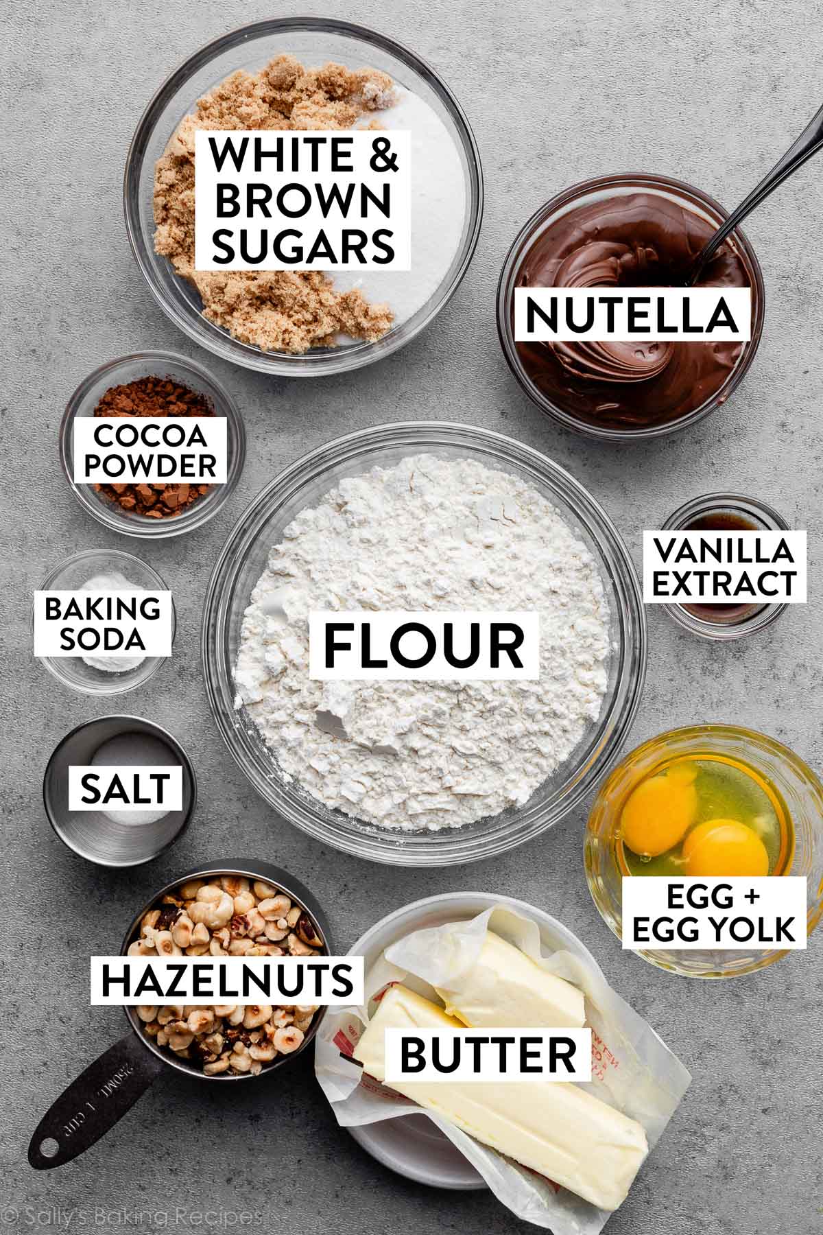 ingredients measured out and on gray surface including flour, Nutella, egg and egg yolk, baking soda, hazelnuts, vanilla extract, and butter.