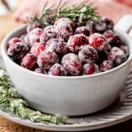 sugared cranberries and rosemary in white bowl on gray plate.