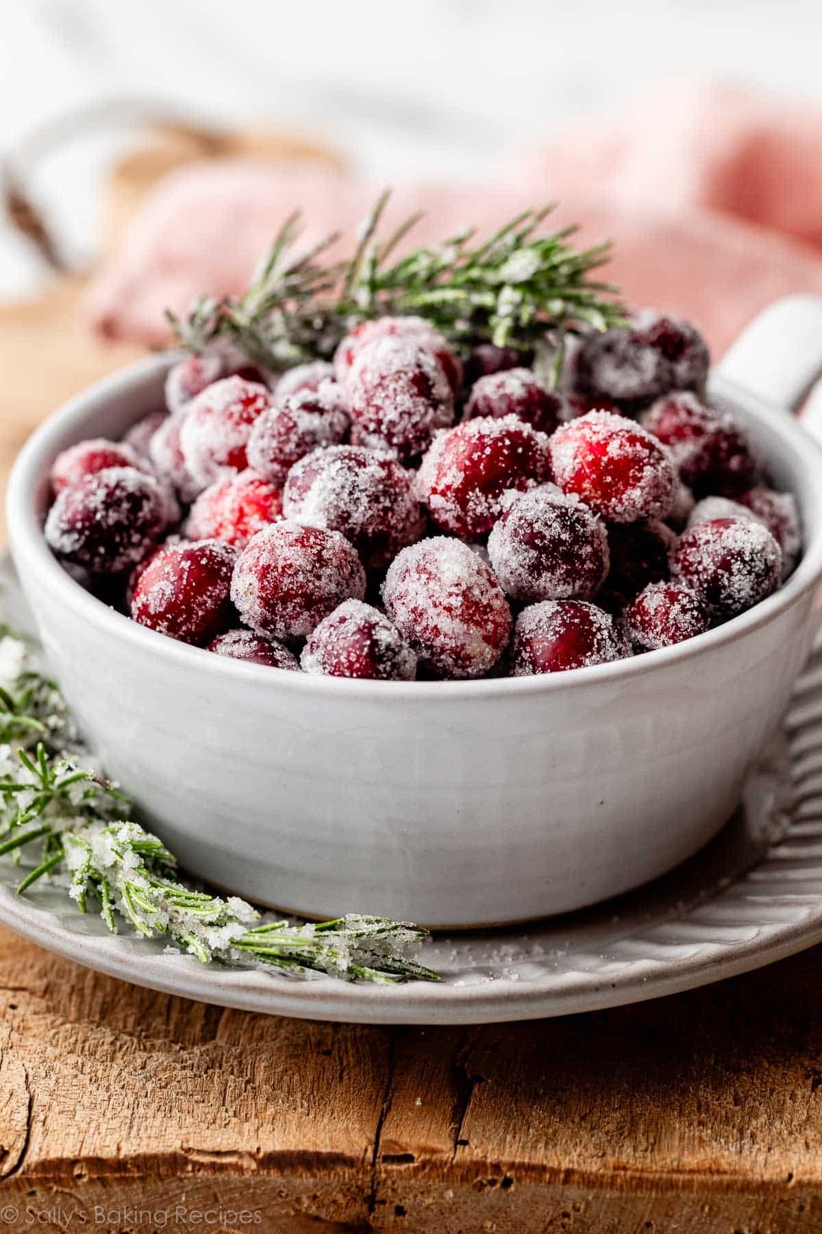sugared cranberries and rosemary in white bowl on gray plate.