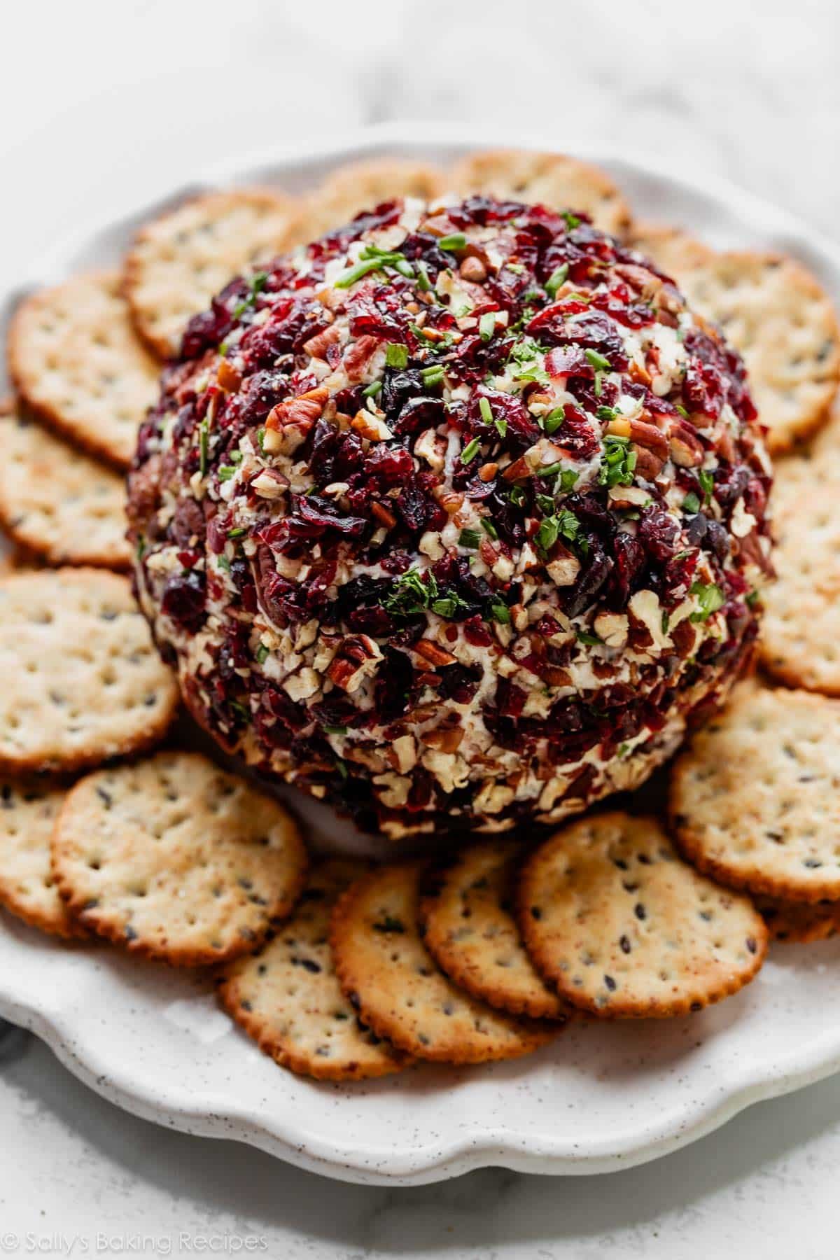 cheese ball coated in pecans, cranberries, and chives on plate with crackers.