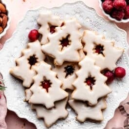 Christmas-tree shaped iced cherry almond linzer cookies on white plate with frozen cherries in frame.