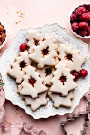 Christmas-tree shaped iced cherry almond linzer cookies on white plate with frozen cherries in frame.