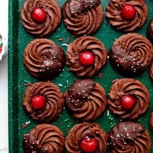 chocolate butter cookies with chocolate and cherries on green baking sheet.