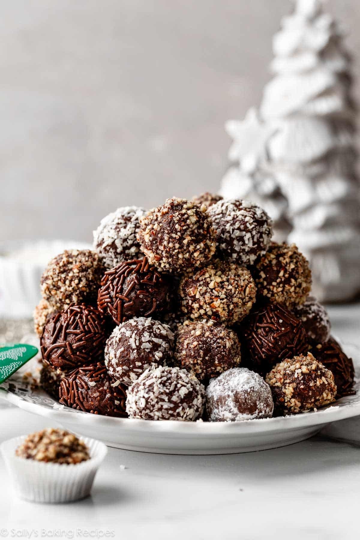 pile of rum balls coated in pecans, coconut, and chocolate sprinkles on white plate.