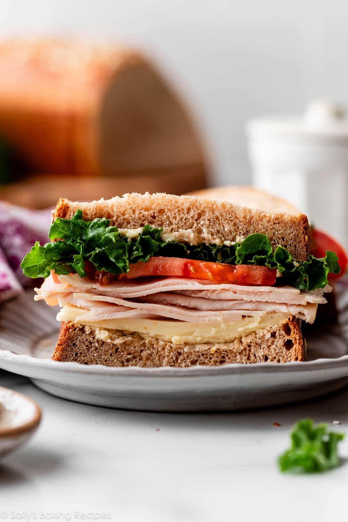 turkey, tomato, lettuce, and cheese sandwich cut open on gray plate with cut loaf of bread in background.
