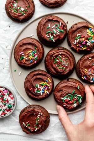 hand grabbing chocolate cookie with chocolate frosting and Christmas sprinkles off plate.