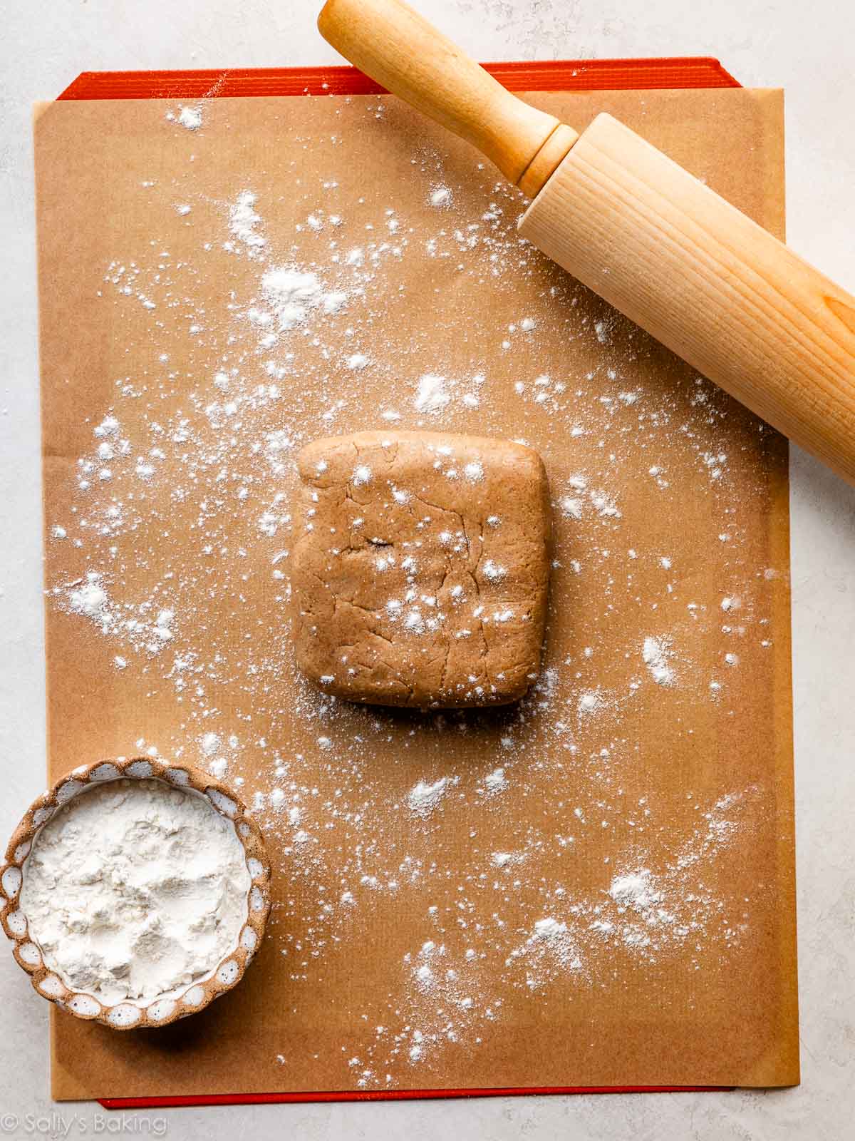 graham cracker dough on a floured piece of parchment paper with a rolling pin and bowl of flour.