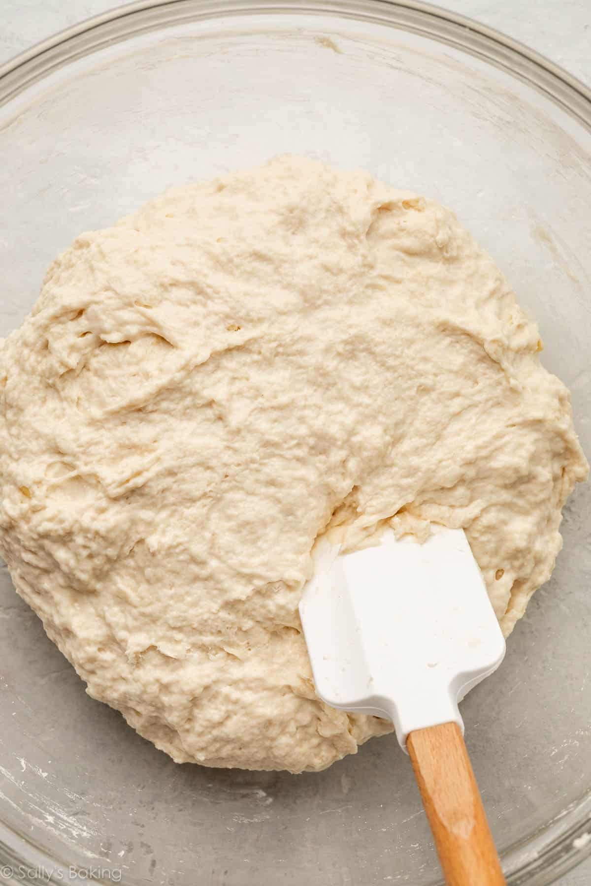 focaccia dough in a glass bowl with a spatula.