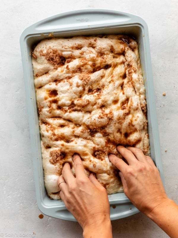 hands dimpling focaccia dough in a baking pan.