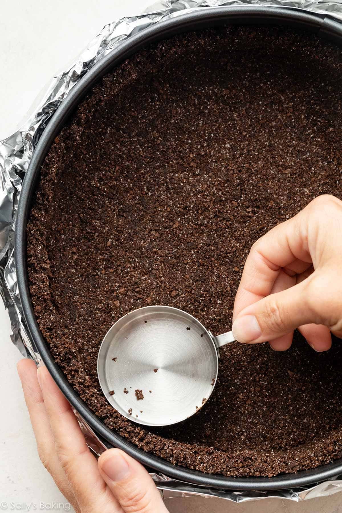hand using a measuring cup to press chocolate graham cracker crumb crust into a springform pan wrapped in foil.